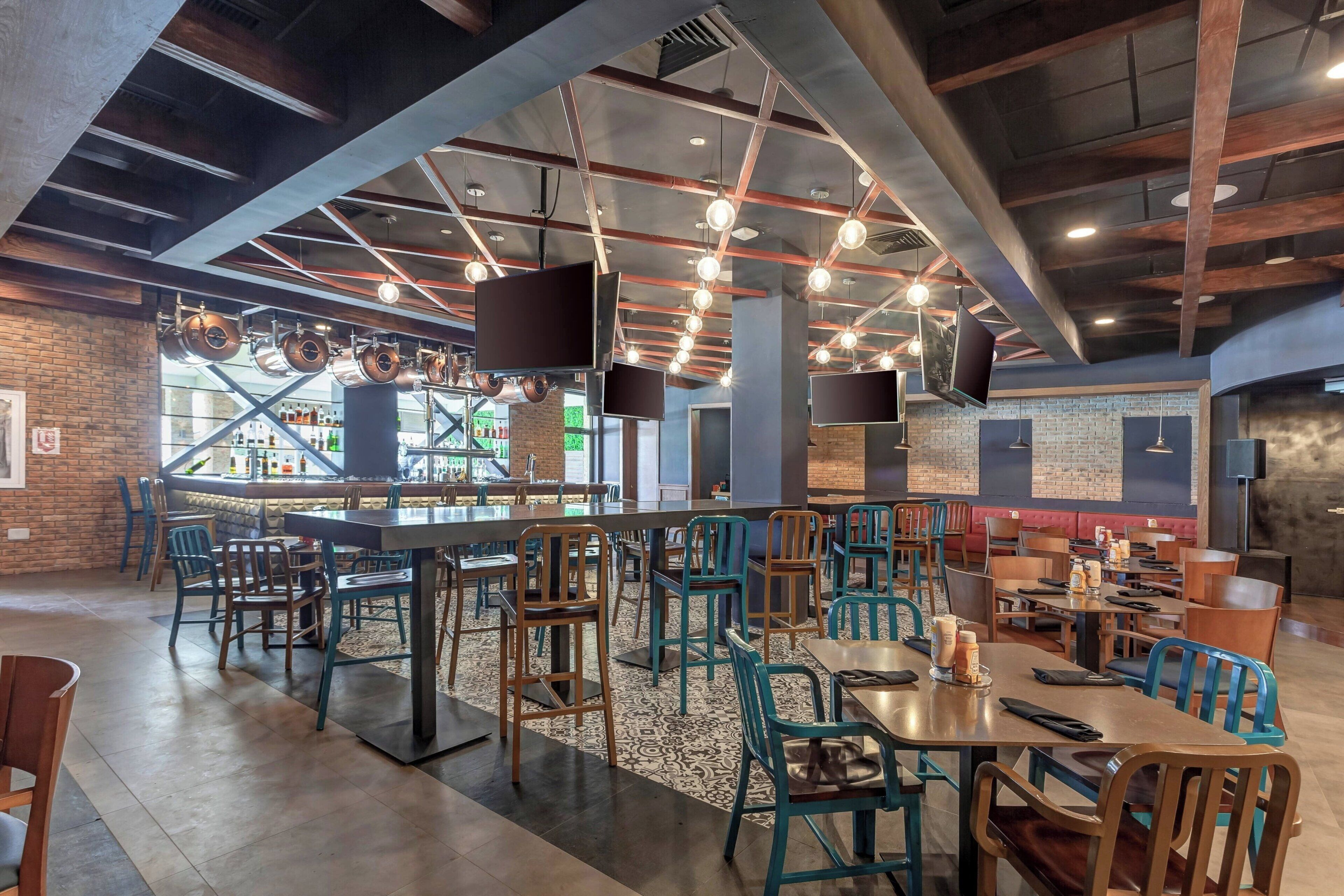 A spacious restaurant with high ceilings, exposed beams, and modern lighting fixtures. Tables and chairs are arranged for dining, and a bar area is visible in the background.