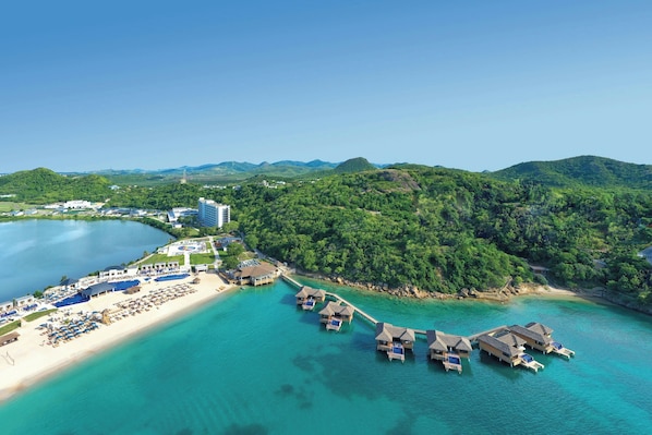 An aerial view of a coastal resort with overwater bungalows, a beach, and lush greenery.