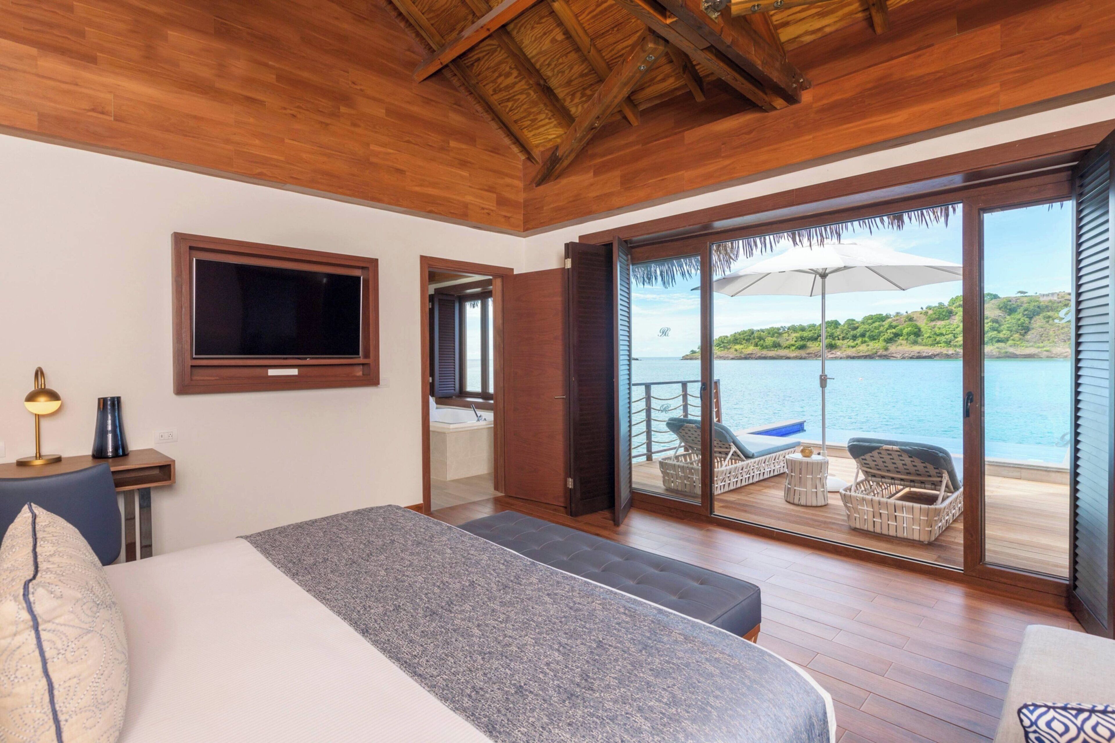 A bedroom with a large bed, a TV, a desk with a lamp, and a view of a balcony with outdoor furniture and a boat.