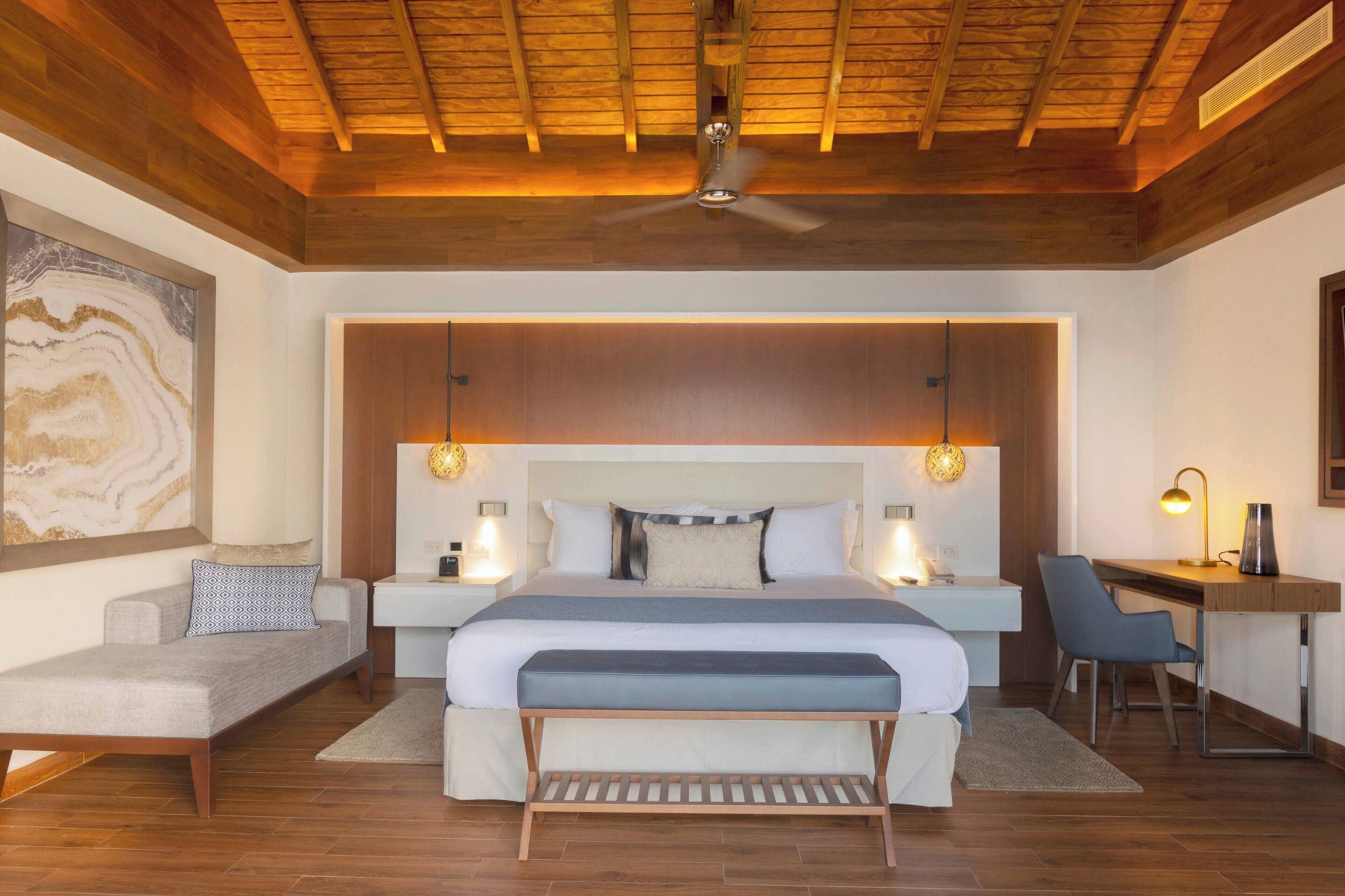 Una camera da letto moderna con soffitto in legno, un letto grande con una panca, comodini con lampade, una scrivania e una sedia.