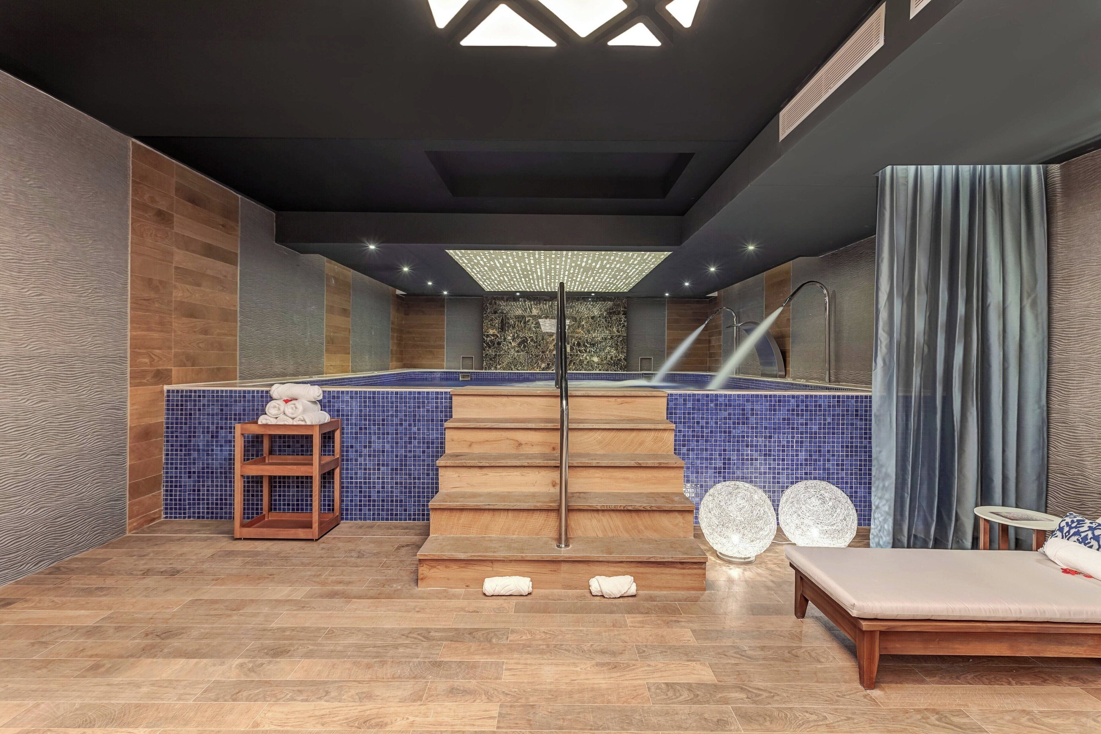 A modern spa area with a wooden staircase leading to a pool, a lounge with a massage table, and a small wooden table with towels.