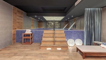A modern spa area with a wooden staircase leading to a pool, a lounge with a massage table, and a small wooden table with towels.