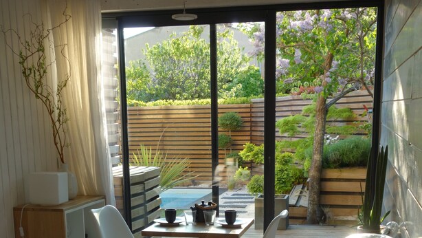 A bedroom with a bed, a table with chairs, a small kitchen area, and a sliding glass door leading to an outdoor garden. - Urbanhouse Cocoon Studio (Toulouse)