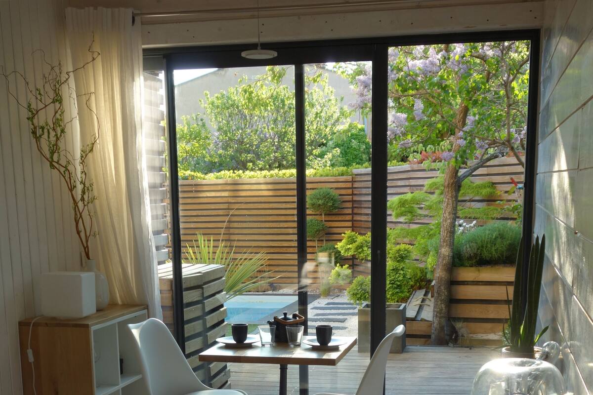 A bedroom with a bed, a table with chairs, a small kitchen area, and a sliding glass door leading to an outdoor garden.