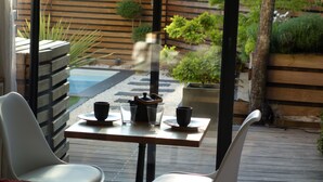 A patio with a table and chairs, a small garden with plants and a wooden fence, and a view of a pool. - Urbanhouse Cocoon Studio (Toulouse)