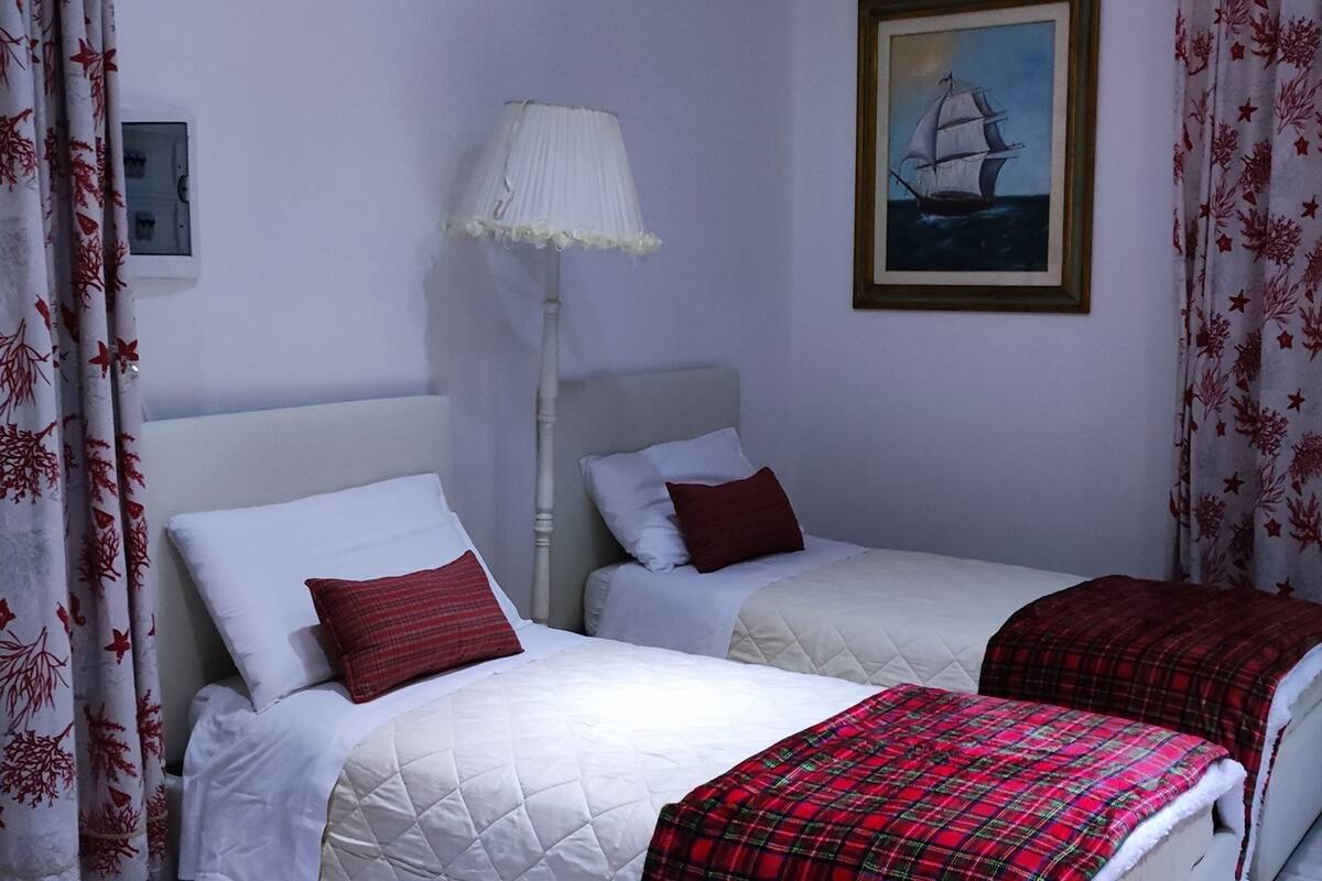Ein Zimmer mit zwei Betten, jeweils mit weißem Kopfteil und roter Bettwäsche. Darüber befindet sich ein Gemälde an der Wand, und eine Stehlampe ist sichtbar.