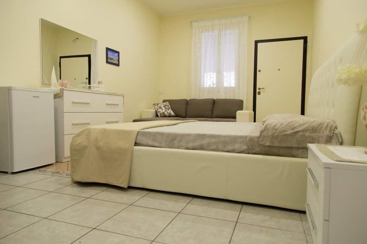 Ein Schlafzimmer mit Bett, Kommode, Spiegel, Fenster mit Vorhängen und einer Tür.