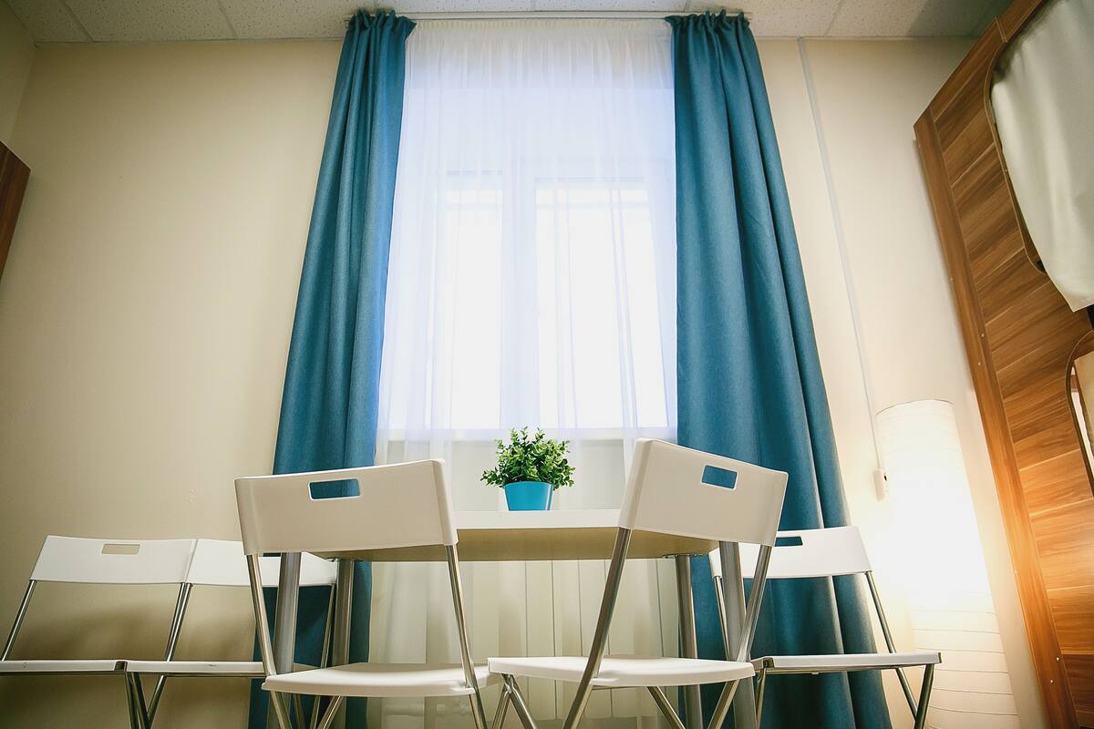 Ein Zimmer mit einem Tisch und zwei Stühlen, einer Zimmerpflanze auf dem Tisch und einem Fenster mit blauen Vorhängen.