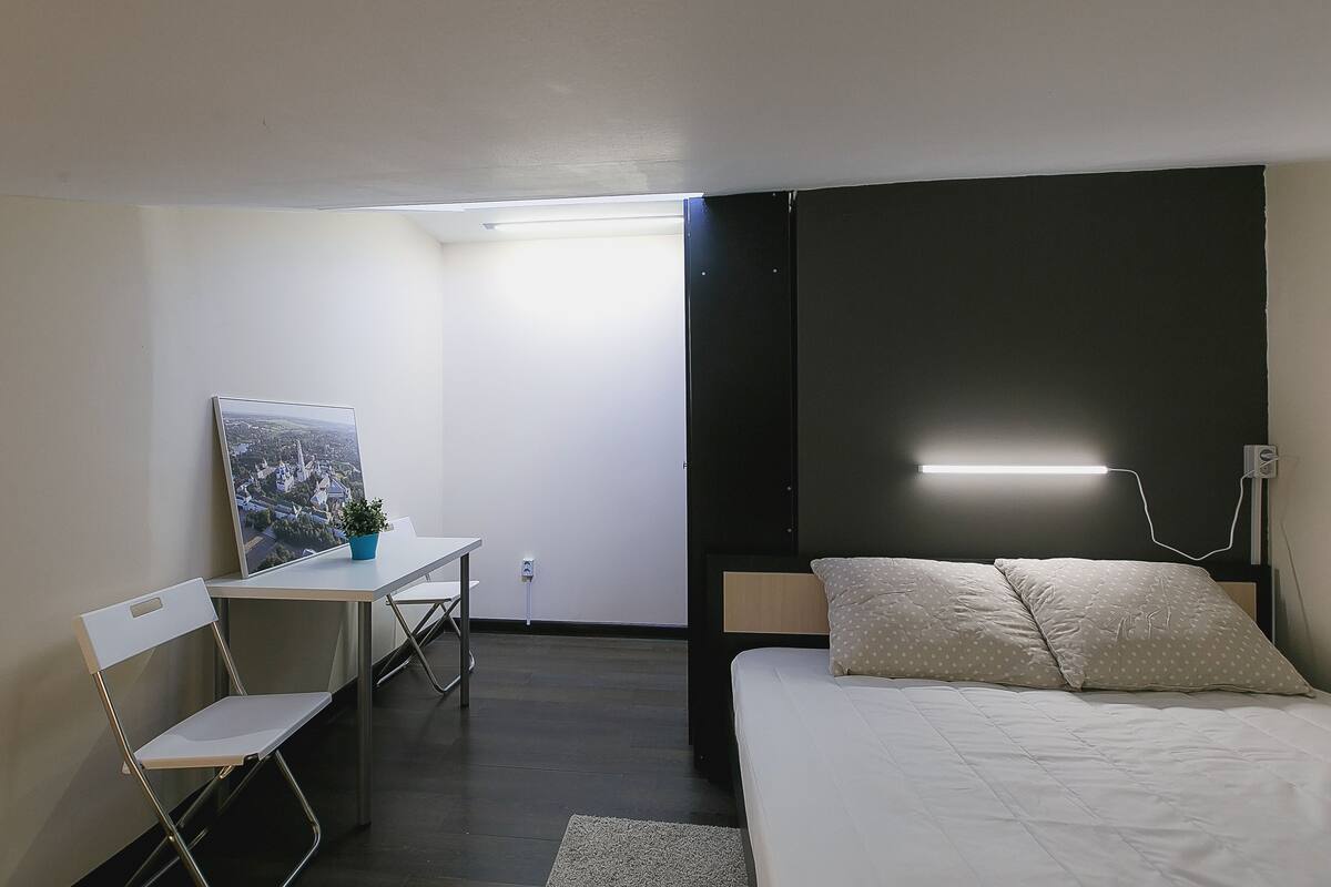 Ein Schlafzimmer mit Bett, Schreibtisch und Stuhl. An der Wand hängt ein Bild und auf dem Schreibtisch steht eine Pflanze.