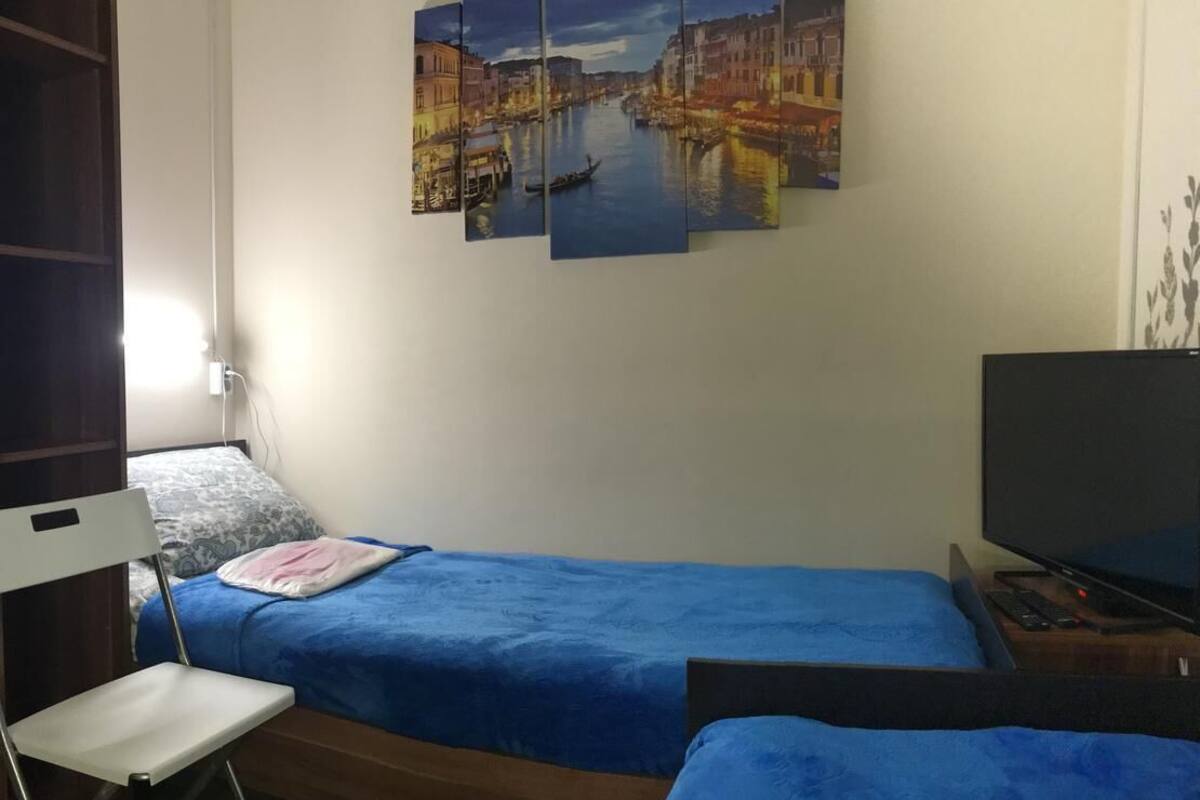Ein kleines Zimmer mit einem Einzelbett, einer blauen Tagesdecke, einem Fernseher auf einem Holzständer und einem an der Wand hängenden Stadtbild.