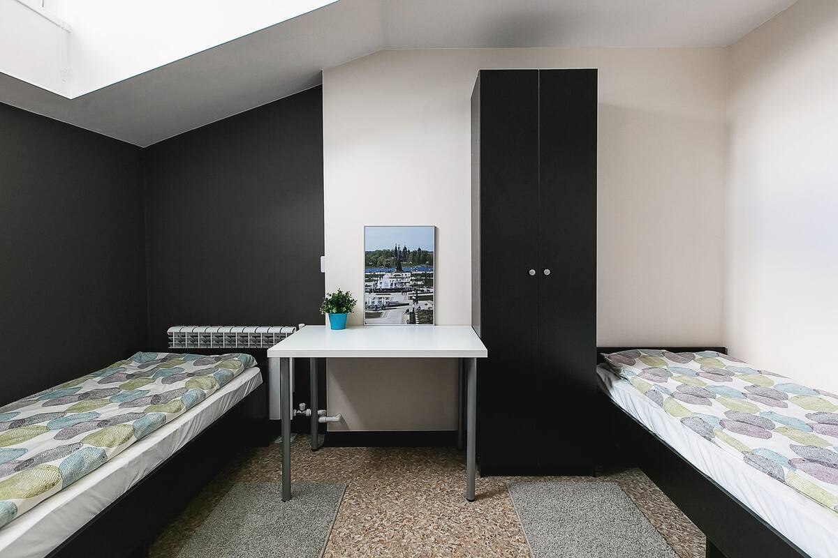 Ein kleiner, moderner Raum mit zwei Einzelbetten, einem Tisch mit einer Pflanze und einem großen Fenster.