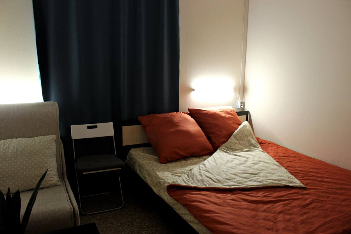 Ein kleines, ordentlich eingerichtetes Schlafzimmer mit einem Bett, einem Sessel und einem Nachttisch.