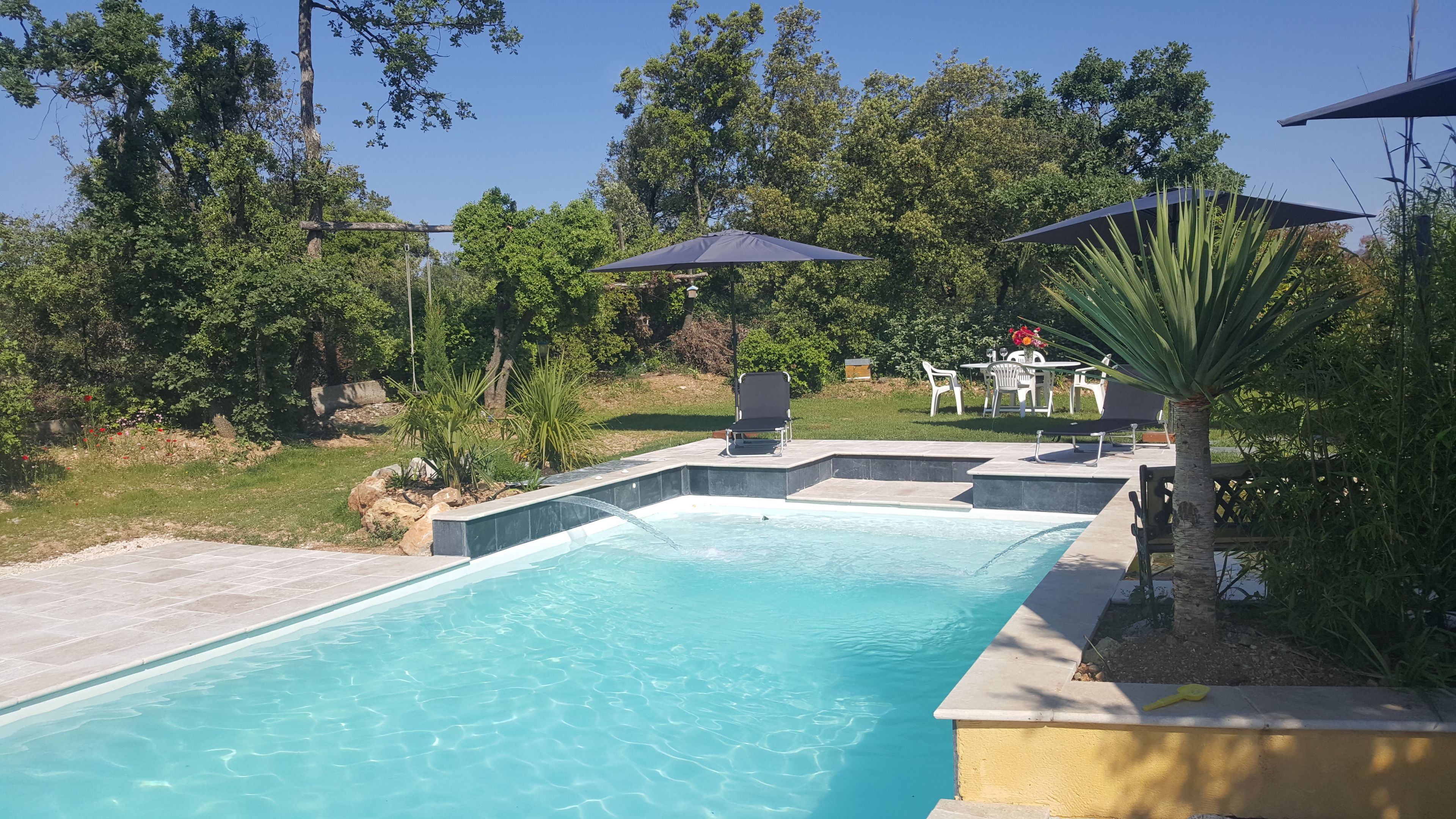 A rectangular swimming pool with a stone edge, surrounded by a patio area with outdoor furniture and umbrellas.