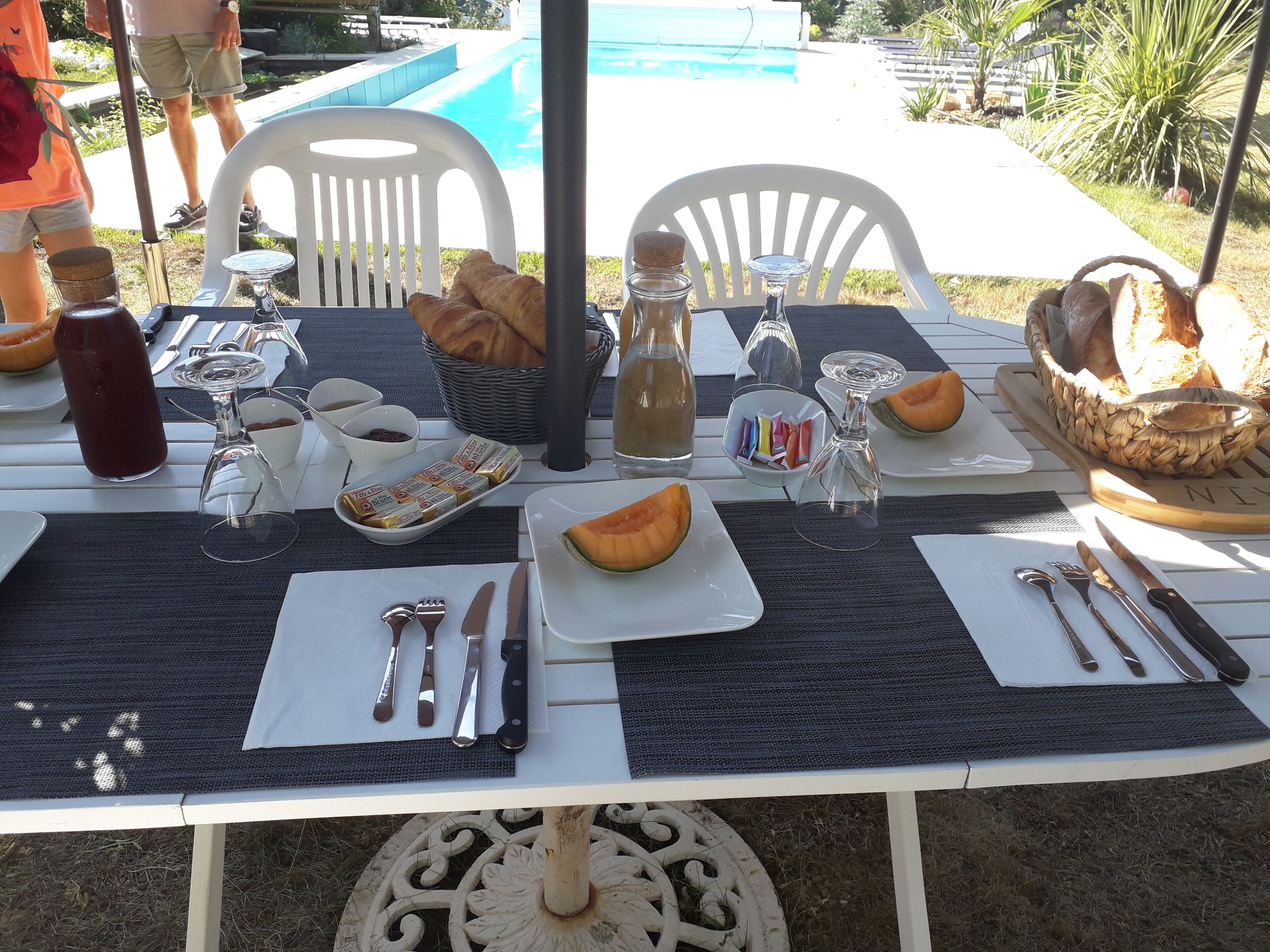 An outdoor dining table set with croissants, bread, a bottle of water, and a plate of melon.