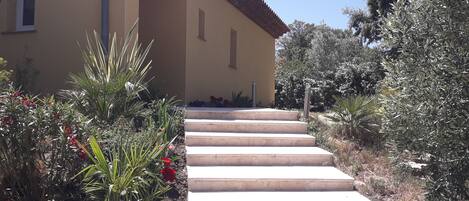 A building with a beige facade and a red-tiled roof, leading up a set of stone steps surrounded by various plants and shrubs.