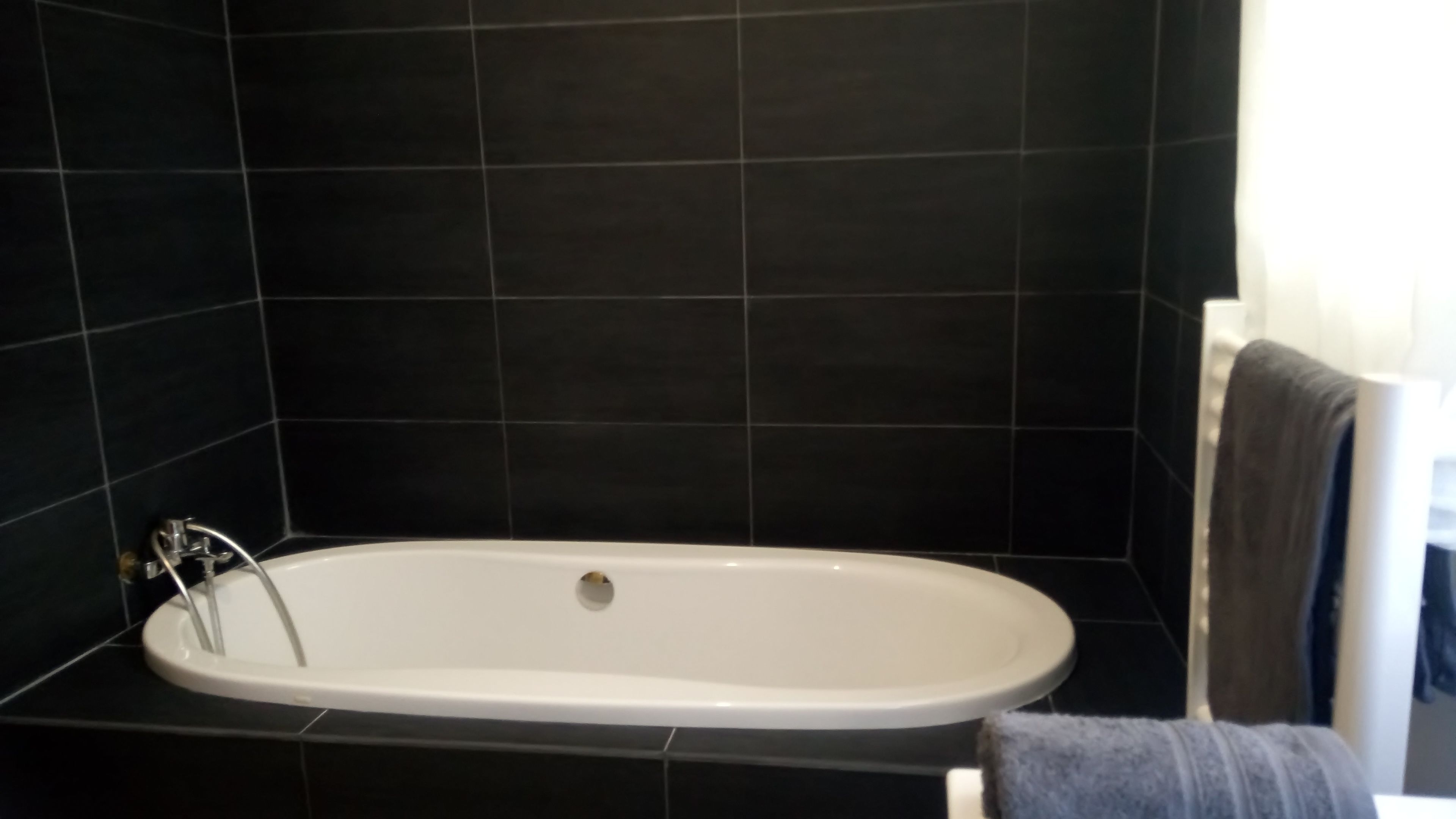 A bathroom with a white bathtub, dark tiled walls, and a towel hanging on a rack.
