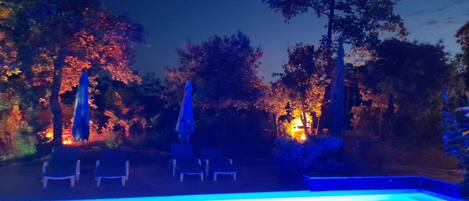 Una piscina iluminada con luces azules, rodeada de árboles y sillas de descanso.