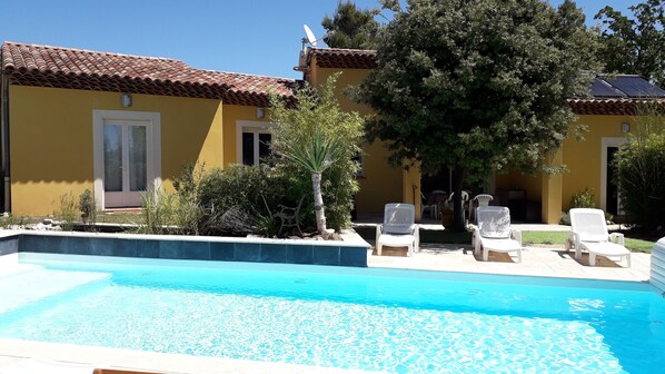 A swimming pool with lounge chairs, a yellow building with a tiled roof, and a tree in the background. - La Villa Provencale (Lorgues)