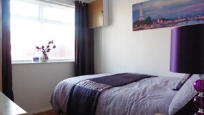 Et soveværelse med seng, et vindue med gardiner, en vase med blomster og et maleri på væggen.