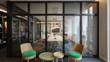 Un intérieur moderne avec un grand mur de verre, deux chaises en bois dotées de coussins verts, une petite table ronde et un espace cuisine visible au fond.