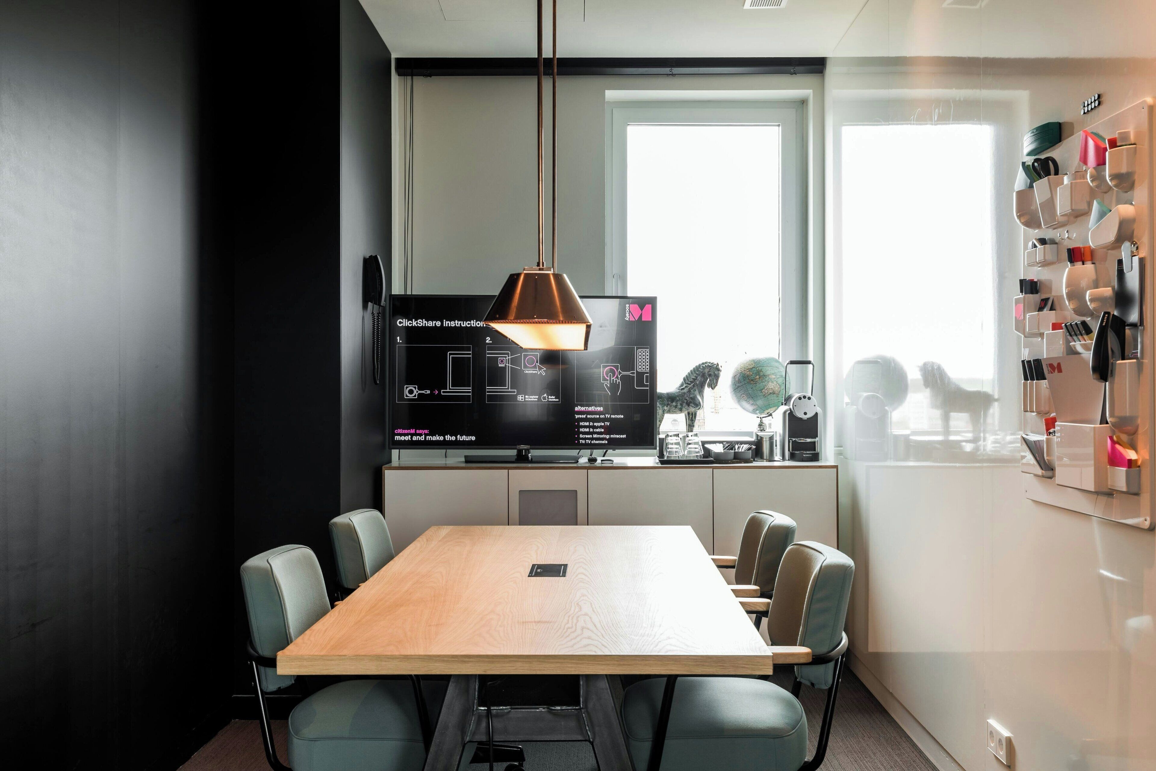 A modern meeting room with a wooden table, six chairs, a large screen displaying instructions, and a wall-mounted shelf with various items.