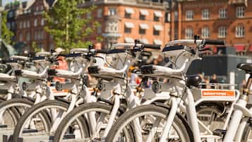 Varias bicicletas estacionadas en un área designada para bicis, con edificios y una bandera al fondo.