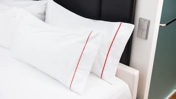 A neatly made bed with white bedding and red piping, a black headboard, and a wall-mounted light switch.