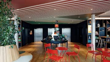 Un vestíbulo moderno con piso de madera, sillas rojas, una mesa central con computadoras portátiles y una planta grande.