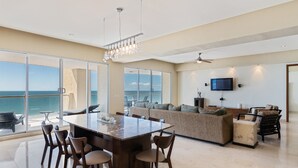 A spacious living area with a large dining table, comfortable seating, a television, and a view of the ocean. - Encanto Living 1103 -3BR by Kivoya (Puerto Peñasco)