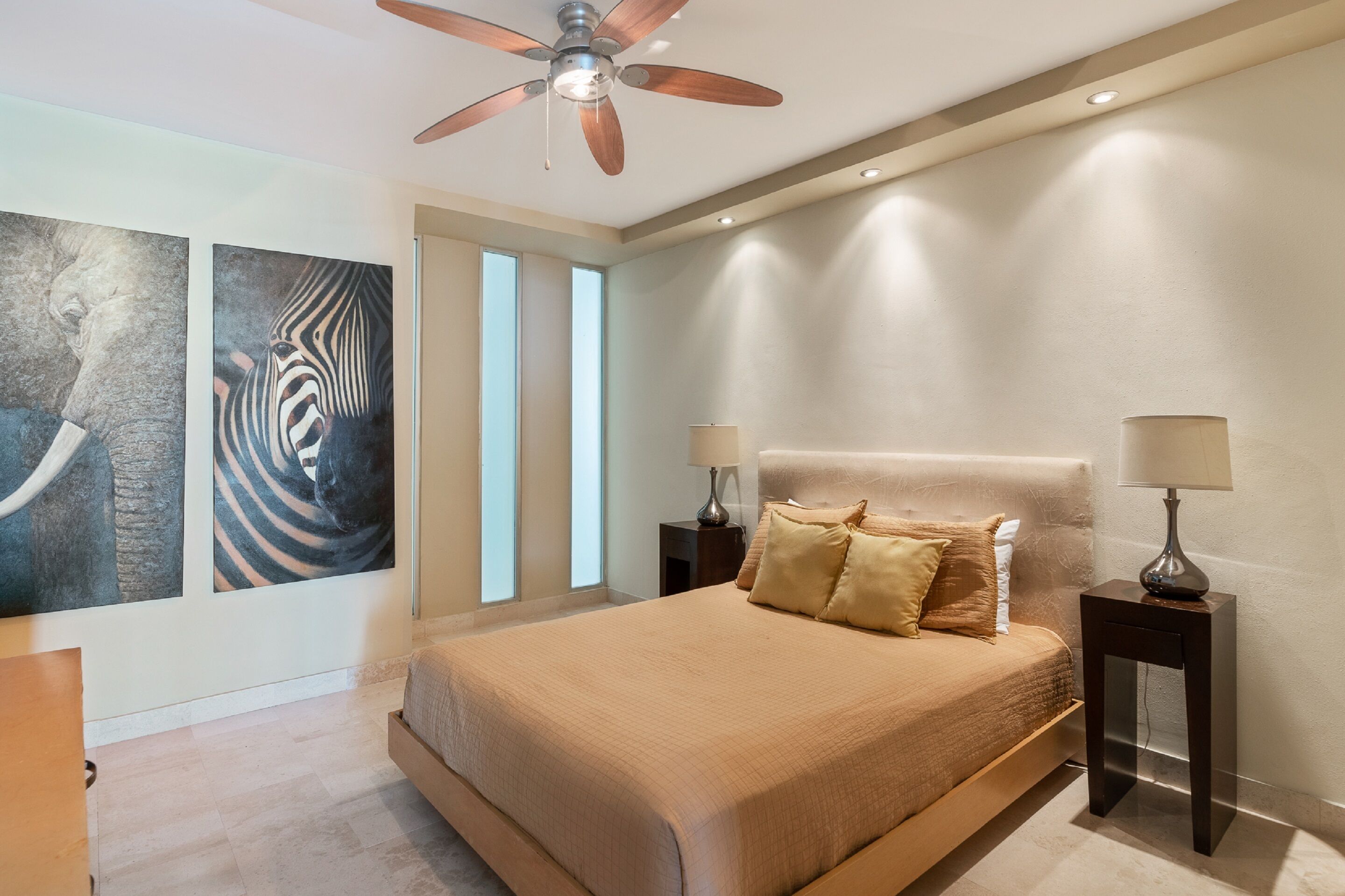 A bedroom with a large bed, two bedside tables with lamps, a ceiling fan, and artwork on the walls.