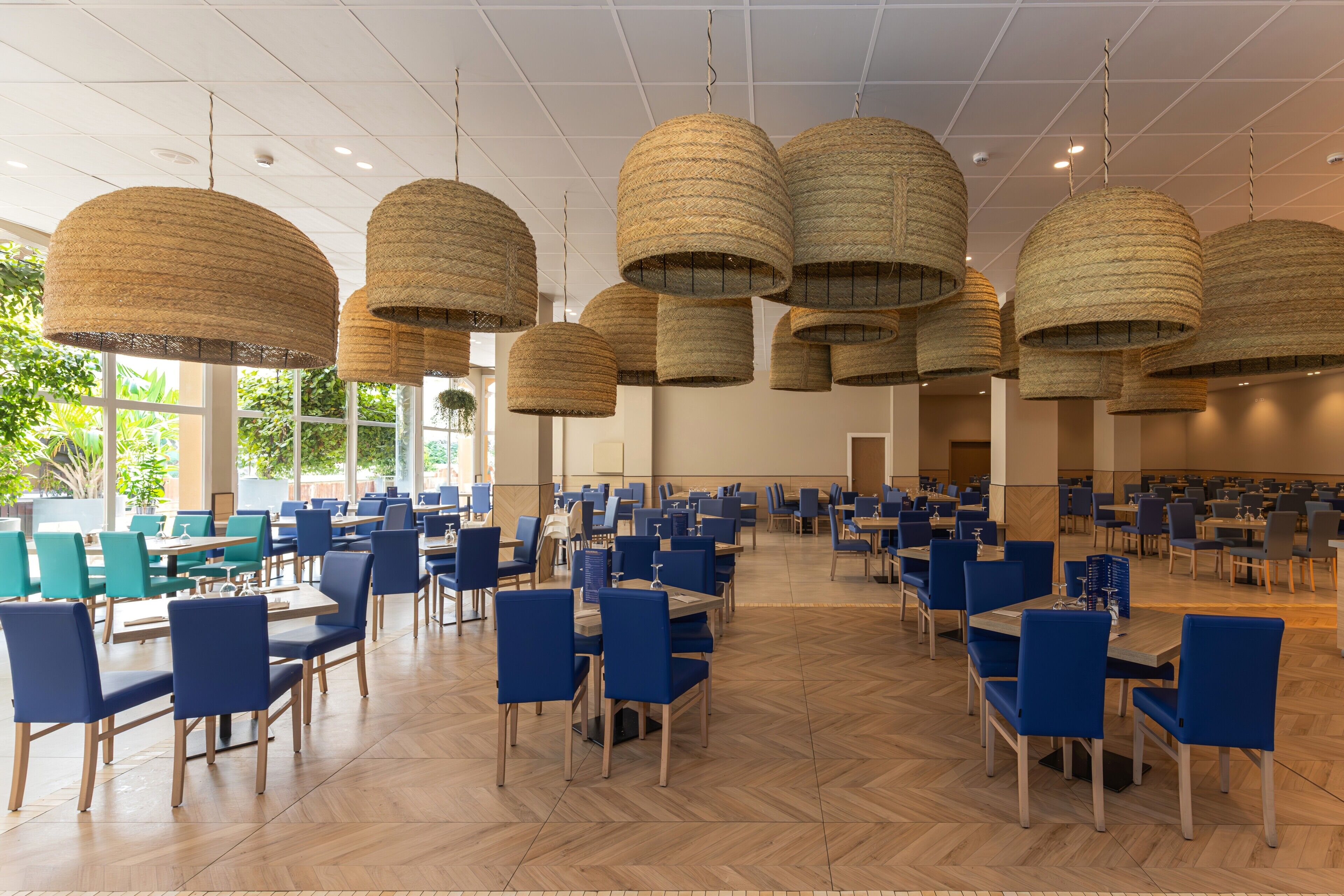 Un vaste espace dédié à la salle à manger, avec de nombreuses tables et chaises, de grands luminaires suspendus en fibres tressées et de grandes fenêtres laissant entrer la lumière naturelle.