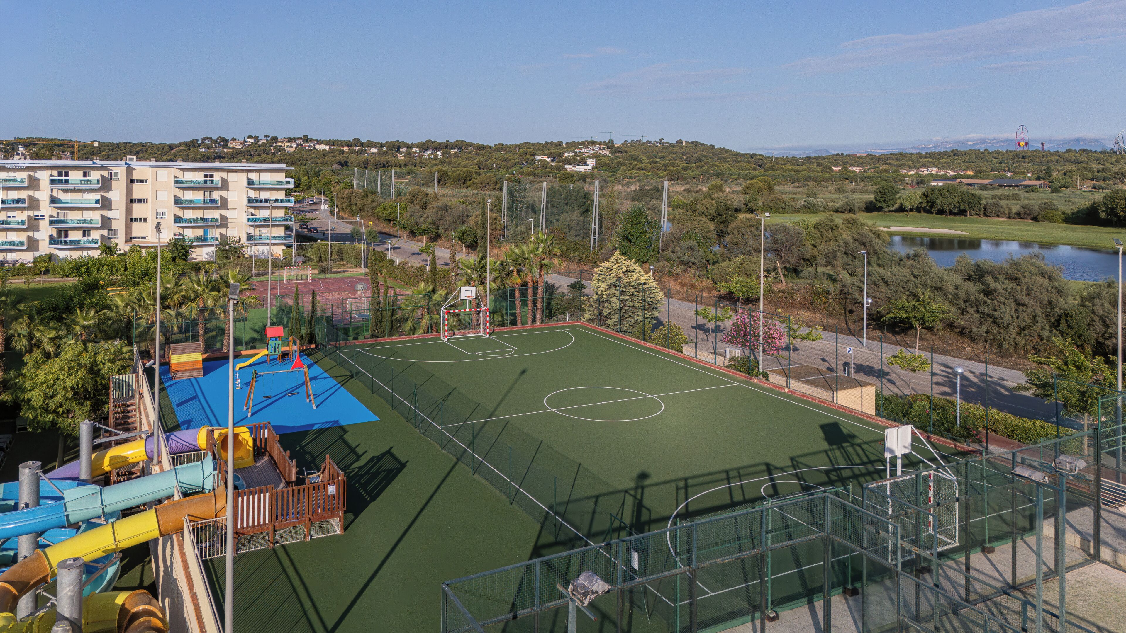 Una cancha de baloncesto con un parque infantil, rodeada de vegetación y una zona residencial.