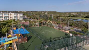 Una cancha de baloncesto con un parque infantil, rodeada de vegetación y una zona residencial.