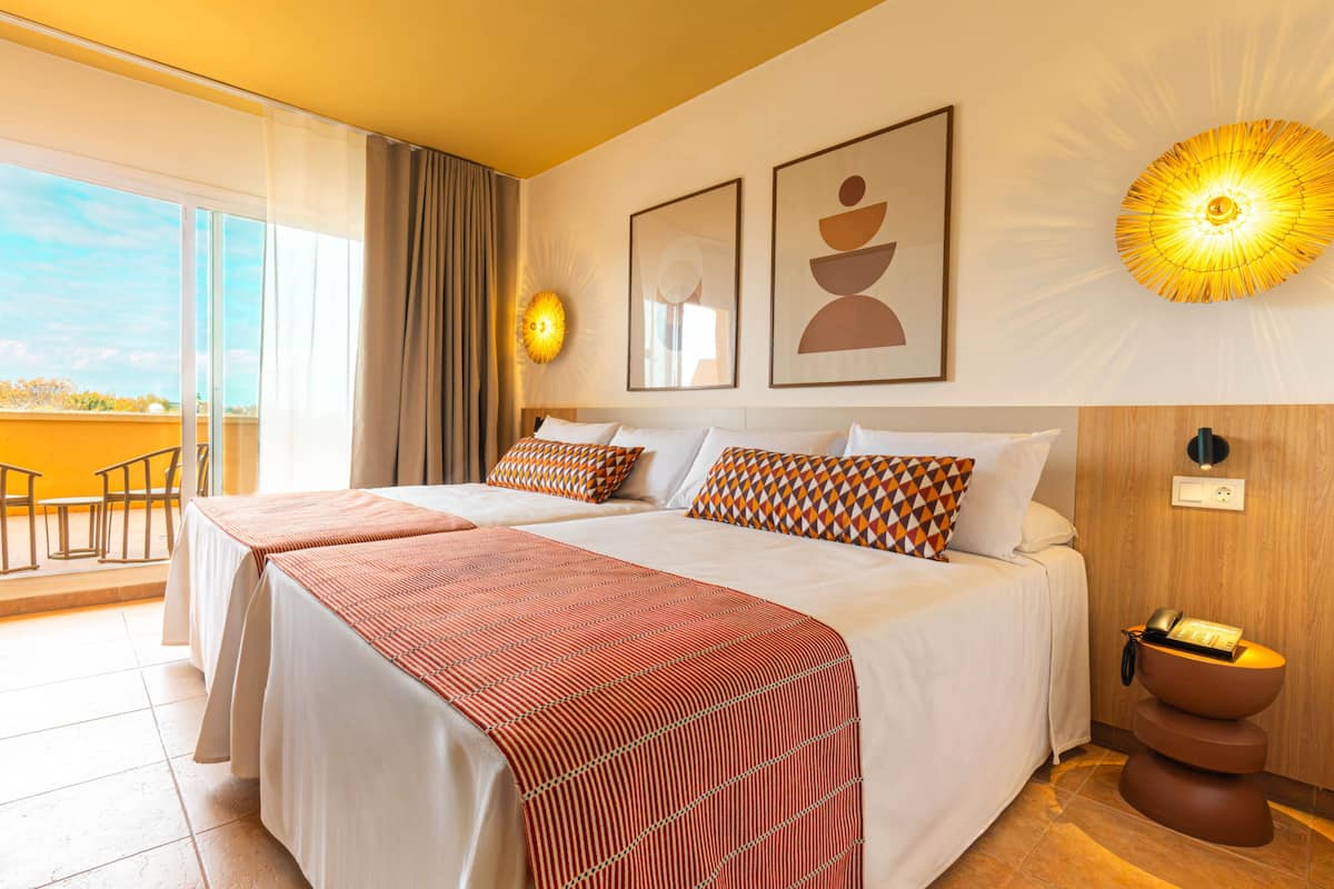 A hotel room with two beds, a balcony, a wall-mounted light fixture, and a wooden side table with a telephone.