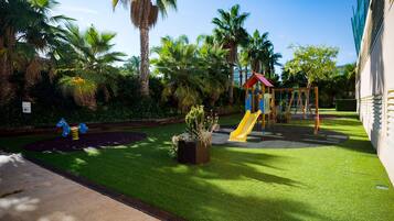 Un parque infantil con tobogán y columpios, rodeado de palmeras y un edificio.