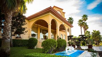 A hotel with a Mediterranean architectural style, featuring a covered patio with arches, a swimming pool, and well-maintained landscaping including palm trees and shrubs.