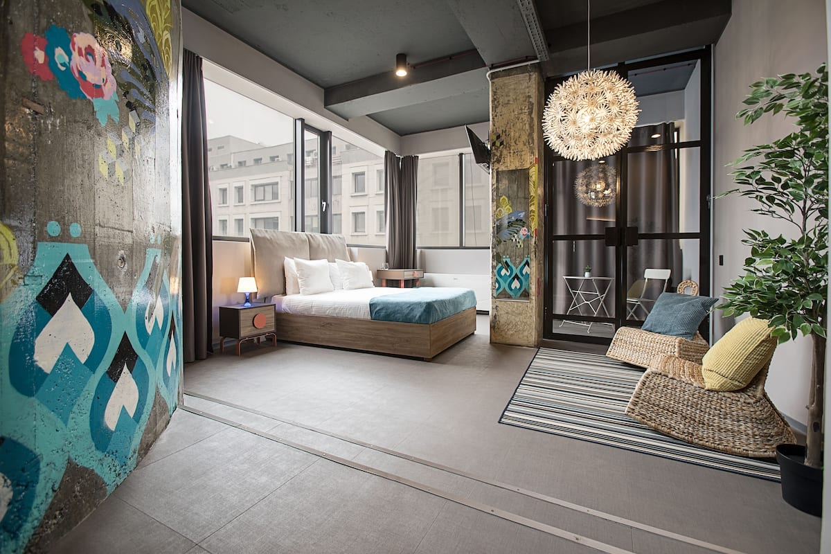 A modern bedroom with a large bed, a bedside table, a lamp, and a view of the city through large windows.