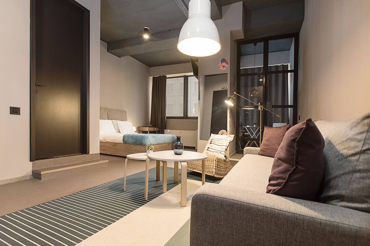 A modern hotel room with a sofa, a small round table, a bed with a blue headboard, and a large window.