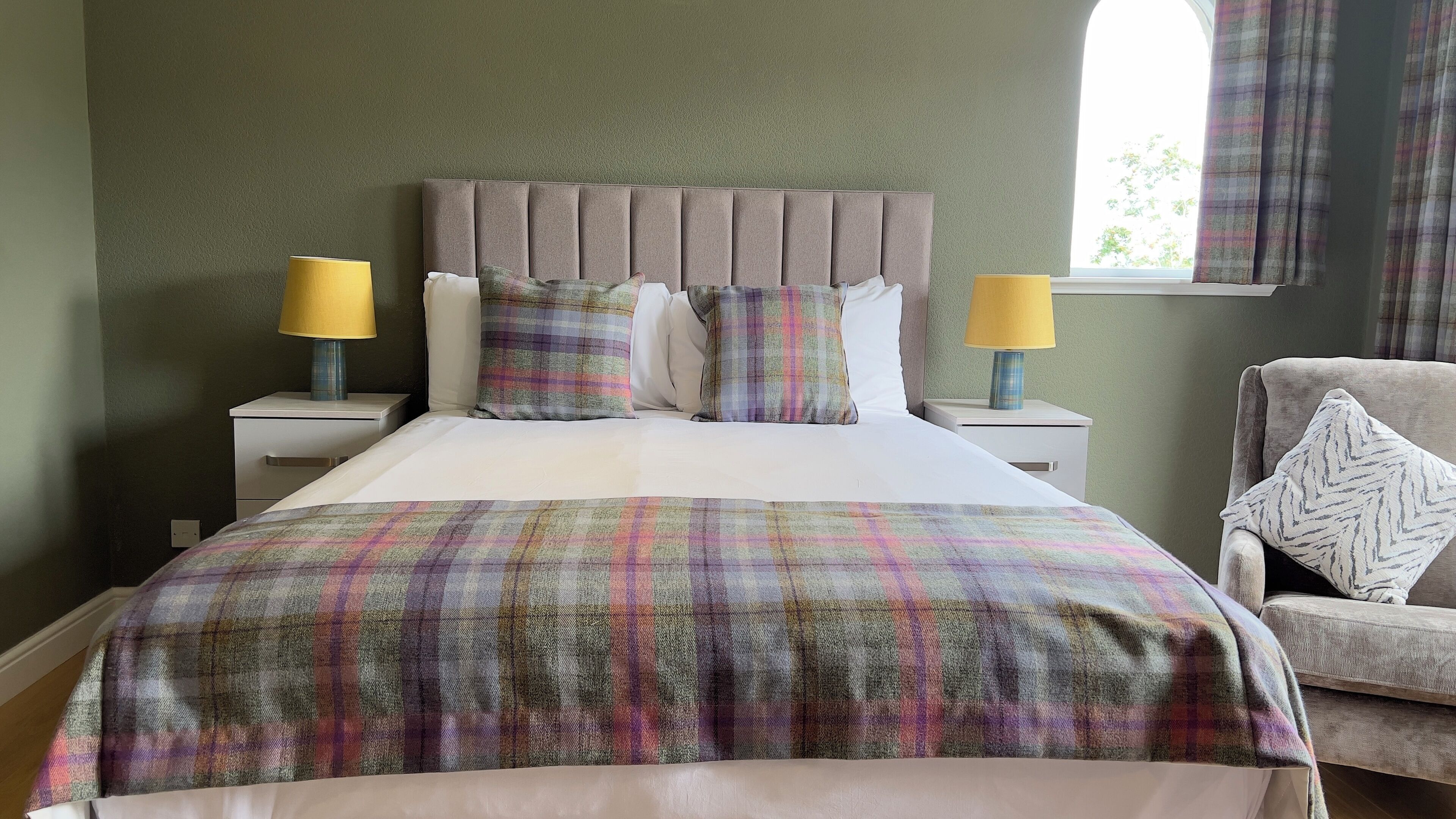 A neatly made bed with a plaid bedspread, two bedside lamps, and a chair with a patterned cushion.