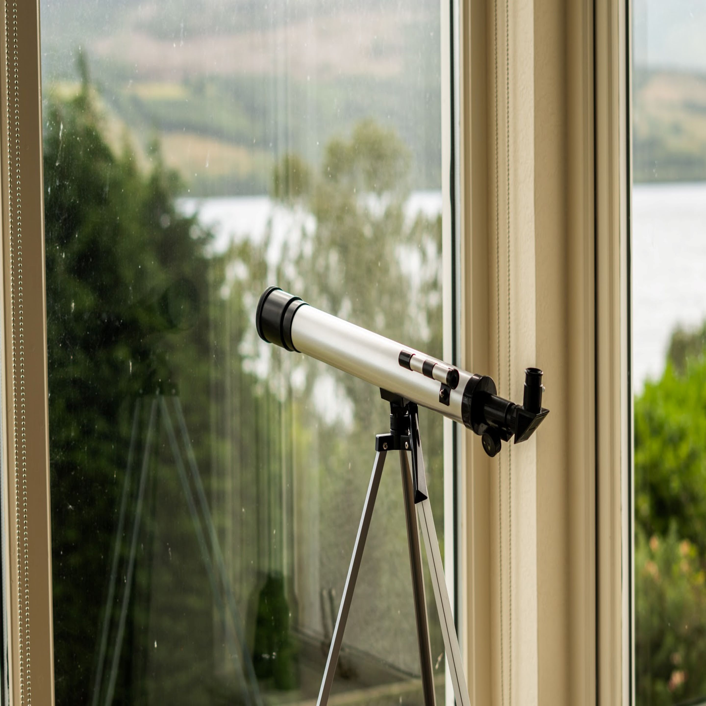 Un telescopio sobre un trípode junto a una ventana con vista a un cuerpo de agua y vegetación.