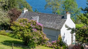 A coastal house with a white exterior, surrounded by lush greenery and flowering plants. - Loch Ness Cottages (Inverness)
