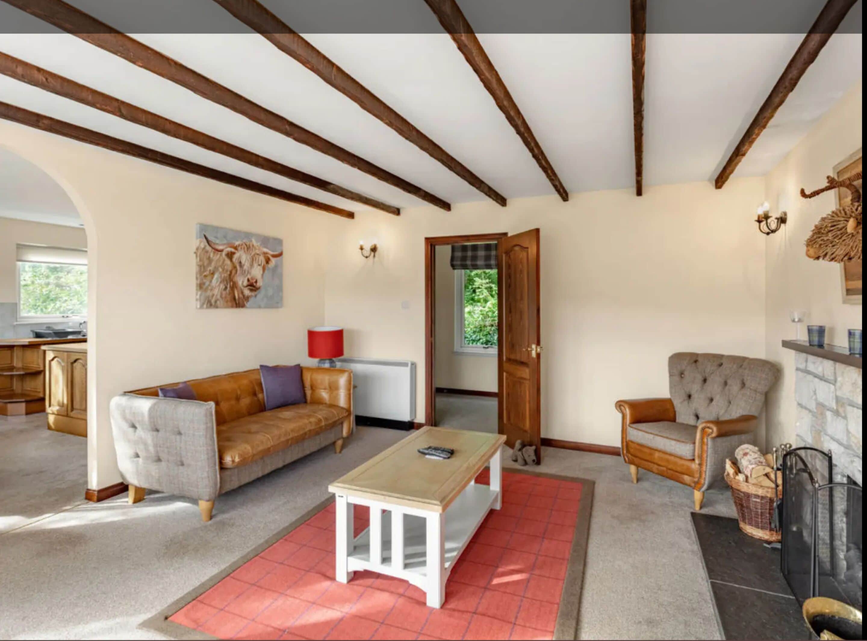 Amplia sala de estar con techo alto, vigas de madera a la vista, un ventanal grande, chimenea y un rincón de estar acogedor con sofá y sillón.