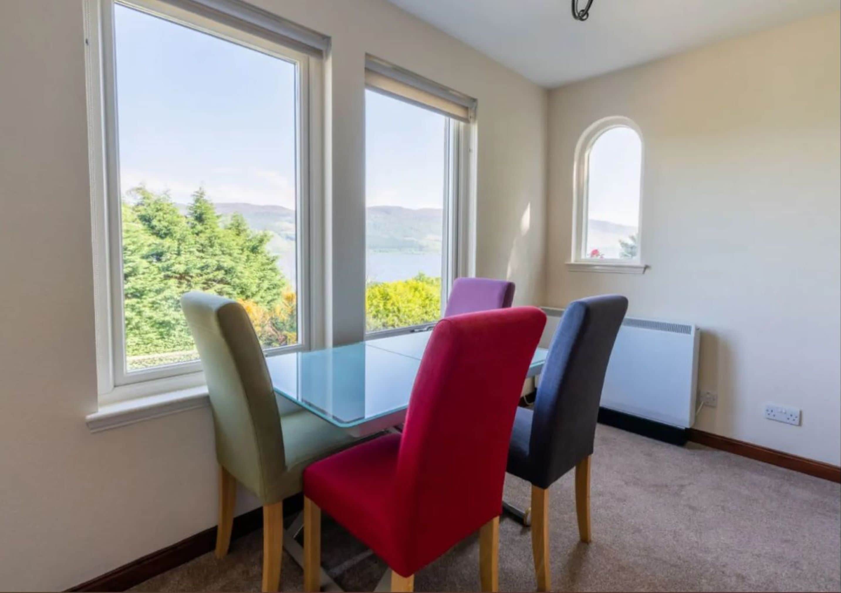 A room with a glass table and four chairs, two windows with a view of greenery and mountains, and a radiator on the wall.
