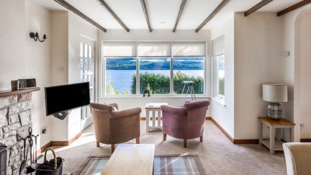 A living room with a large window offering a view of the sea, a fireplace, and a flat-screen TV. - Loch Ness Cottages (Inverness)