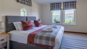 A neatly made bed with a grey headboard, white and red pillows, and a plaid blanket. There are two windows with plaid curtains, and a view of greenery outside. - Loch Ness Cottages (Inverness)