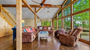A spacious living room with wooden floors, a ceiling fan, large windows, and a staircase. - Lakefront “Treehouse”: Private Retreat in the Pines! (Point Blank)