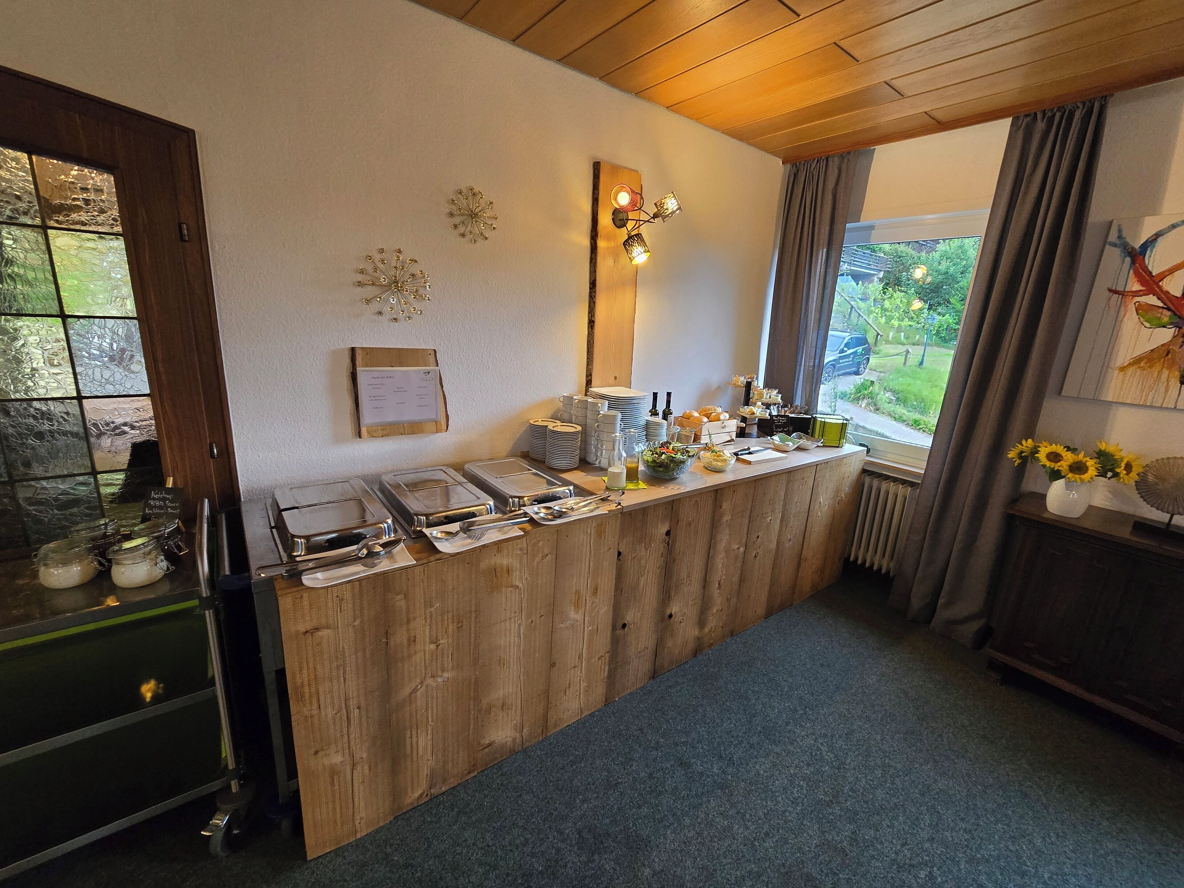 A breakfast bar with various food items and utensils, a wooden counter, and a view of the outdoors through a window.