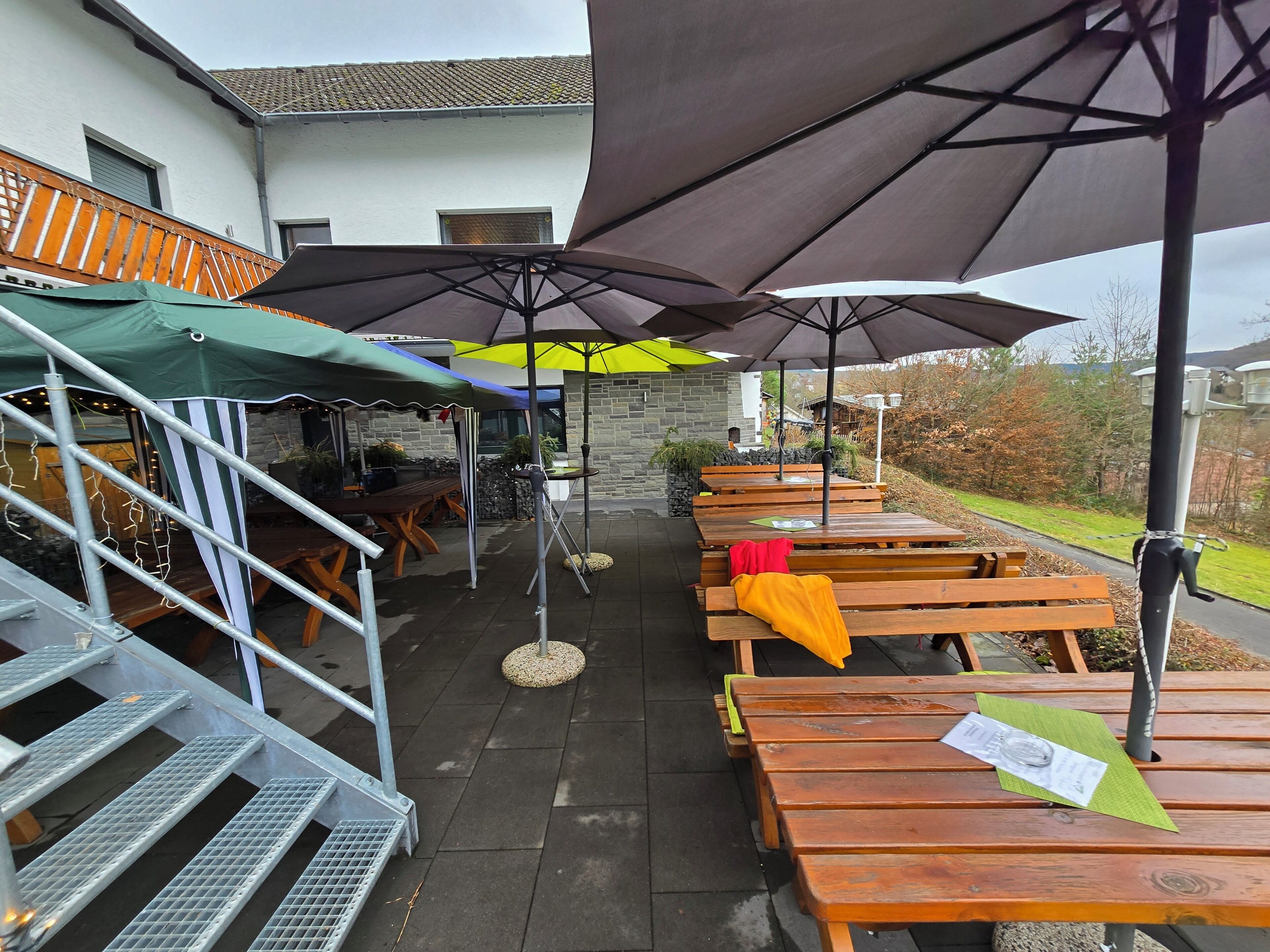 An outdoor patio with wooden tables and umbrellas, a staircase, and a stone wall.