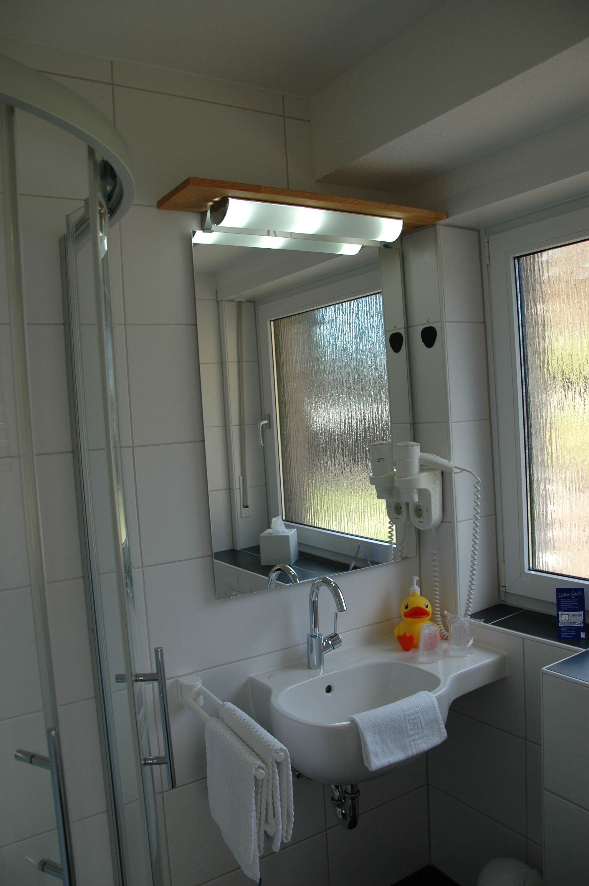 Et badeværelse med glasvæg omkring bruseniche, vask med spejl og et vindue med satin glas.