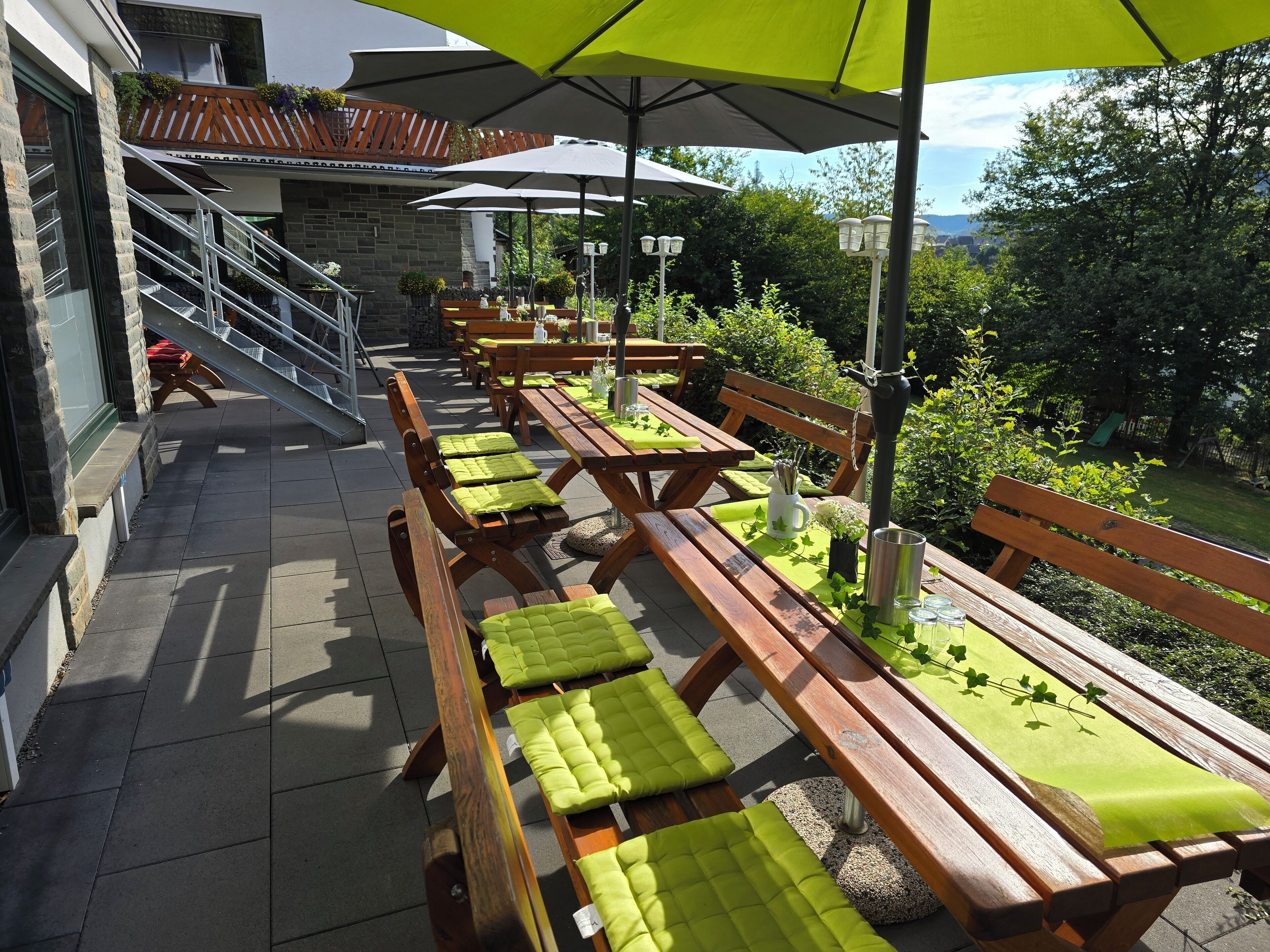 Un patio al aire libre con bancos y mesas de madera, algunas con cojines verdes, y sombrillas que dan sombra.